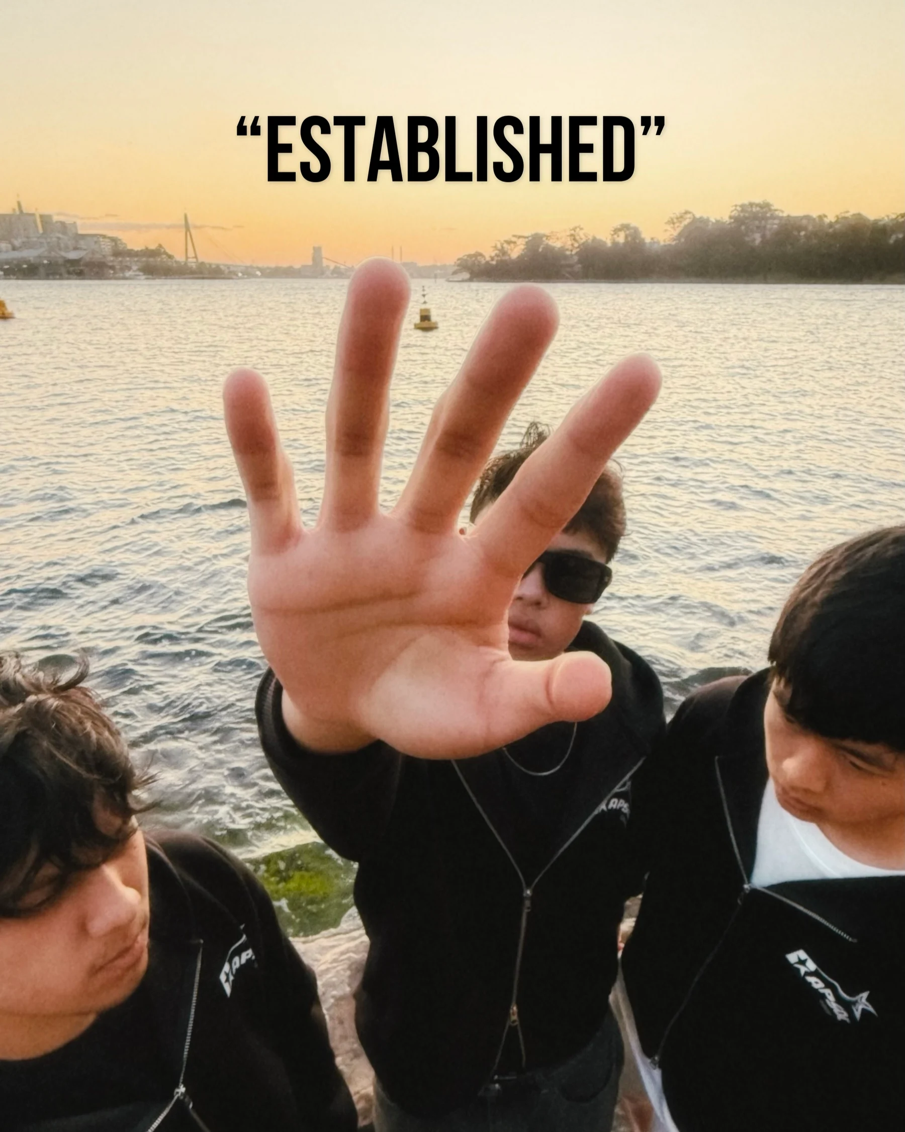 Three RAPSOC members in the black double-zip hoodie by the water at sunset, one holding a palm up to the camera. Overlaid text reads “Established”.